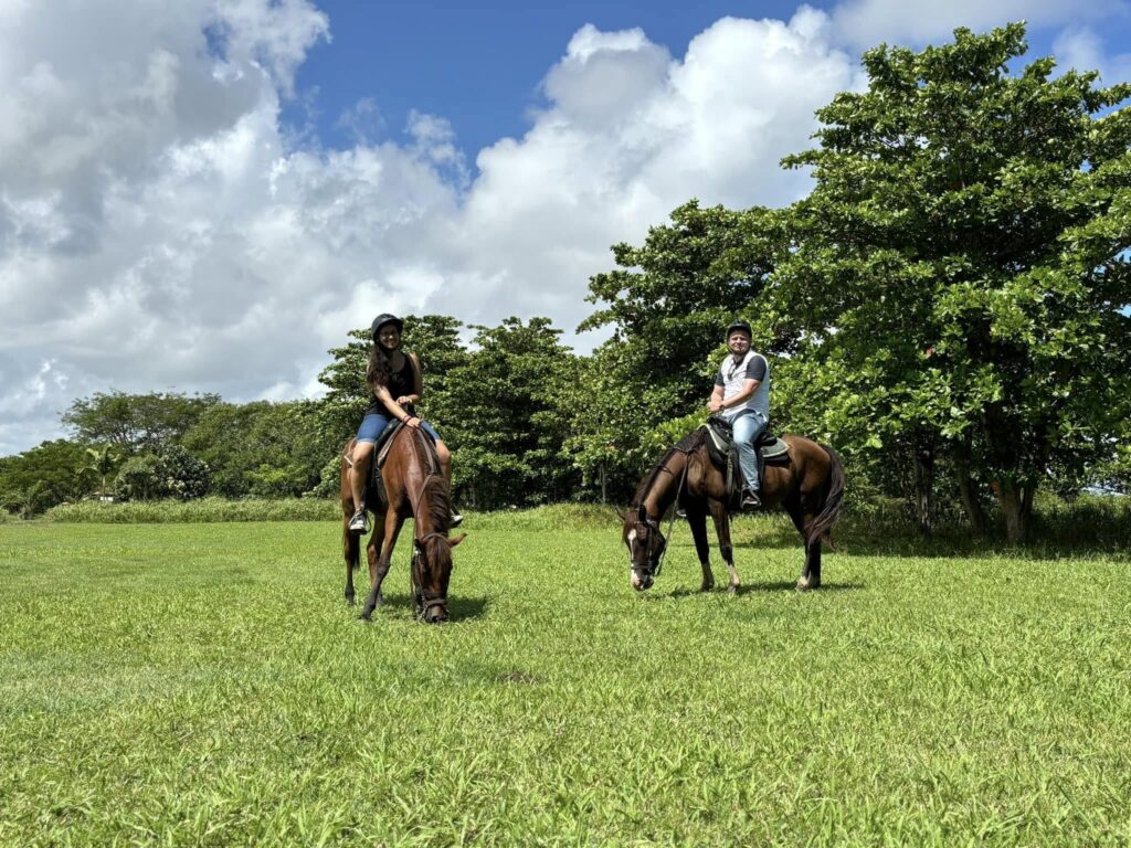 Couple horseback riding through open grassy fields at Hacienda Campo Rico in Puerto Rico, surrounded by lush green trees under a bright cloudy sky.