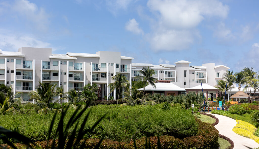 Exterior view of Dreams Jade Resort in Mexico, showcasing white modern buildings surrounded by lush tropical greenery and palm trees under a bright blue sky.
