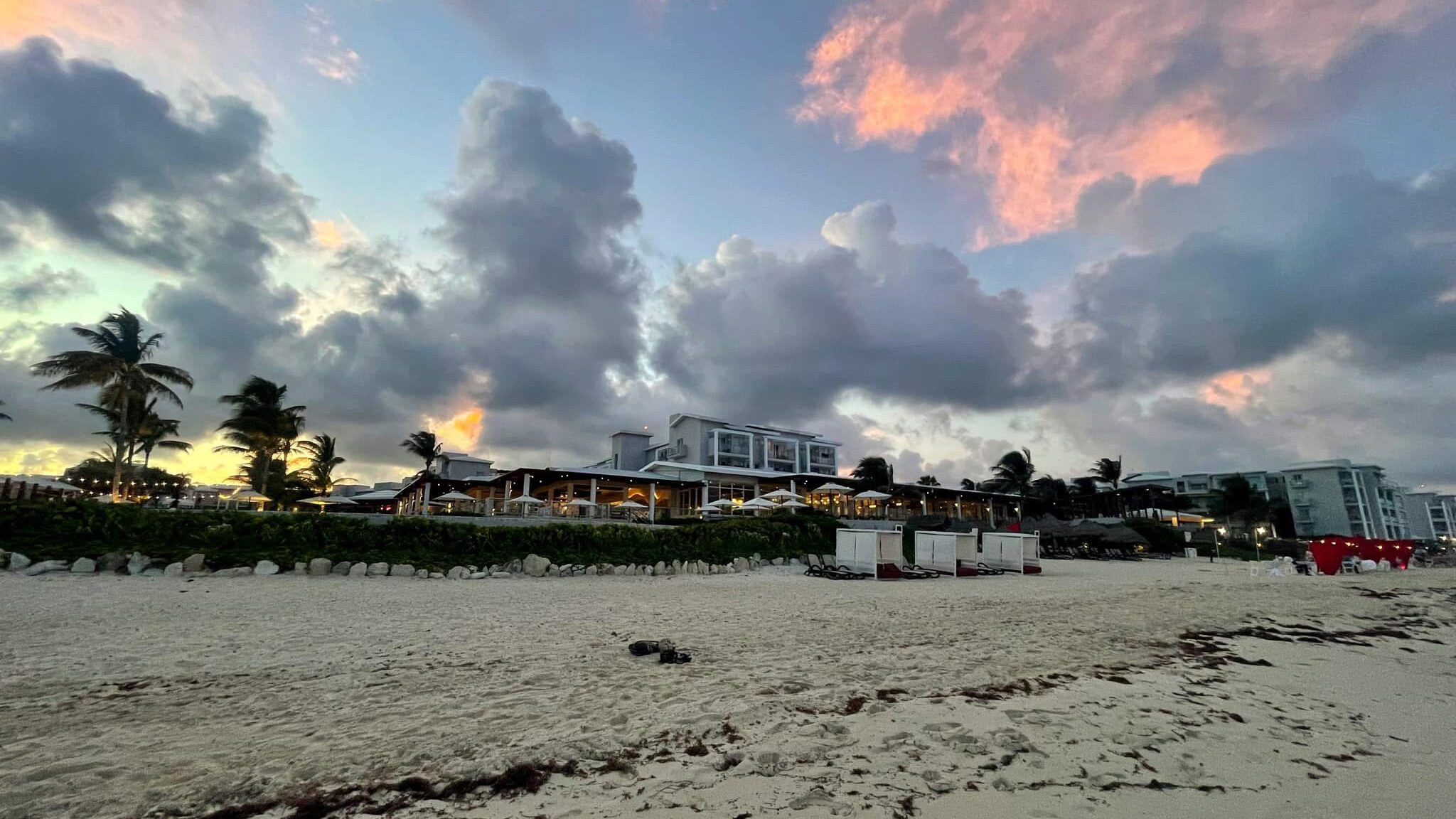 Sunset view of Dreams Jade Resort in Mexico, with dramatic clouds, palm trees, and a peaceful sandy beach lined with cabanas and lounge chairs.