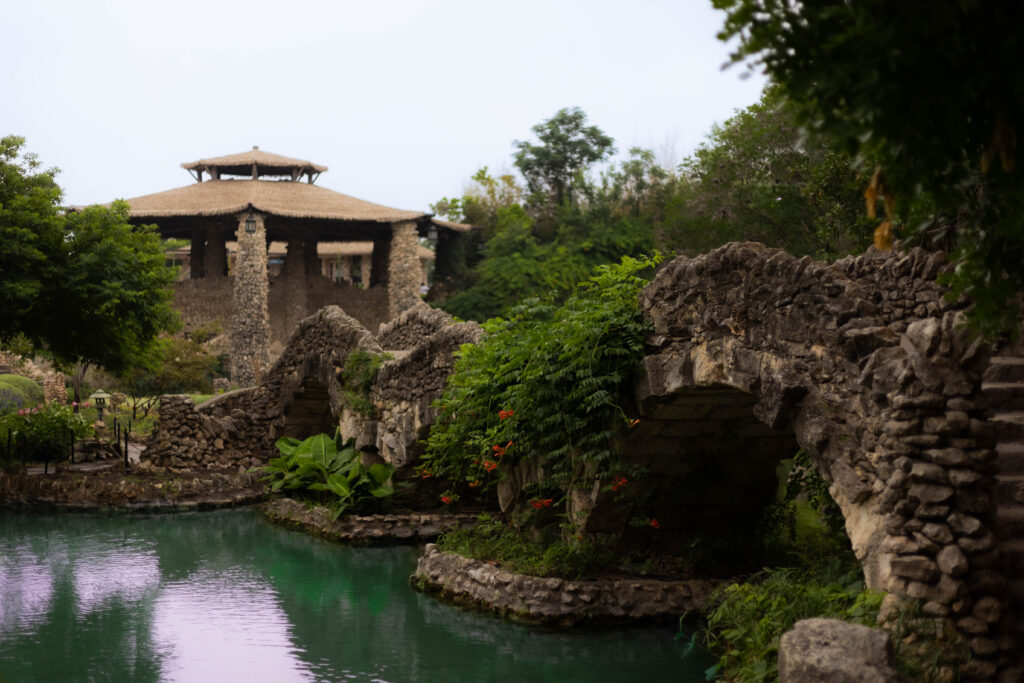 Stone bridge over turquoise water surrounded by lush greenery at a historic garden, with a rustic stone pavilion in the background.
